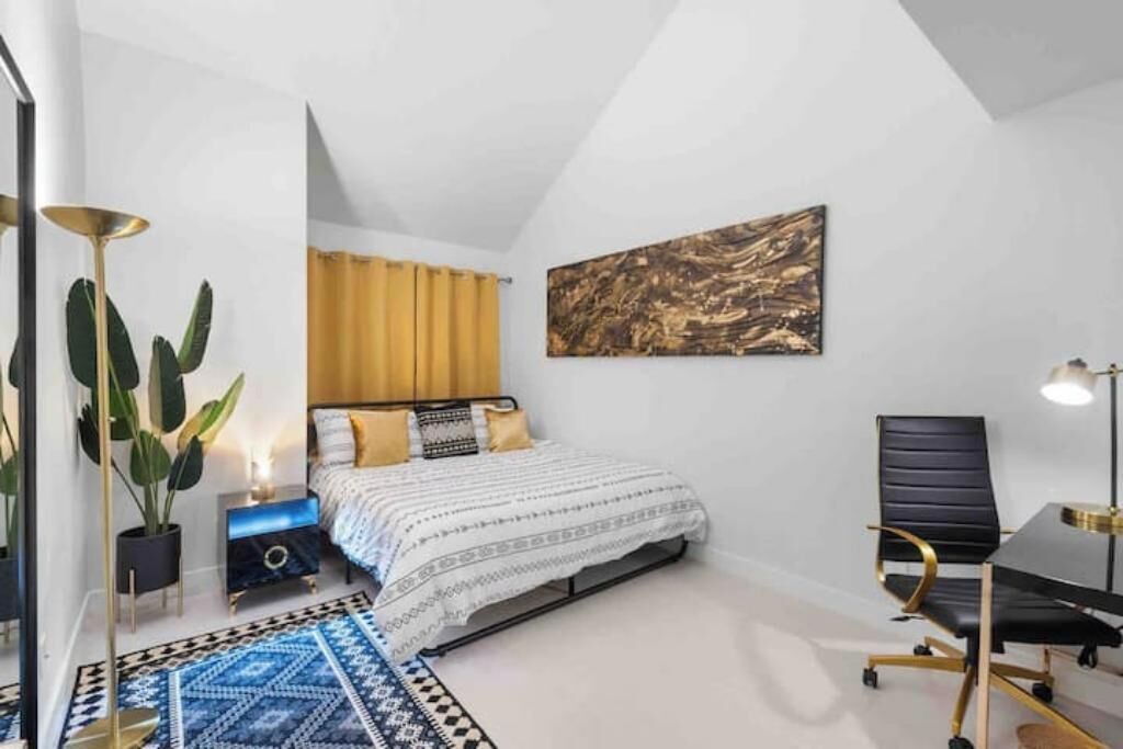 Une chambre à coucher comprenant un lit, une table de chevet, une lampe, une chaise et un grand tableau abstrait accroché au mur.