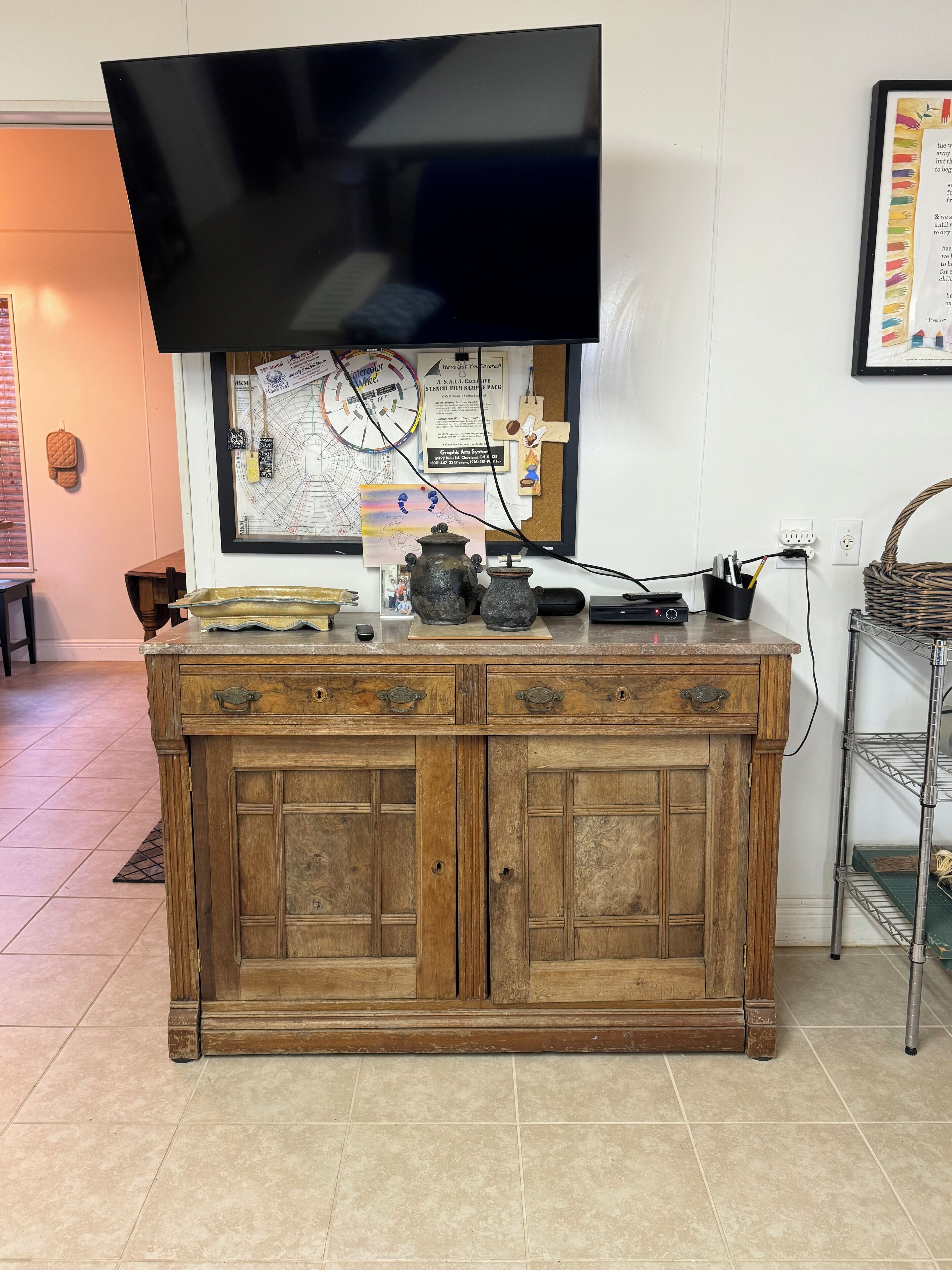 Un mueble de madera con un televisor de pantalla plana montado encima, un cartel enmarcado en la pared y una cesta de mimbre sobre una estantería metálica.