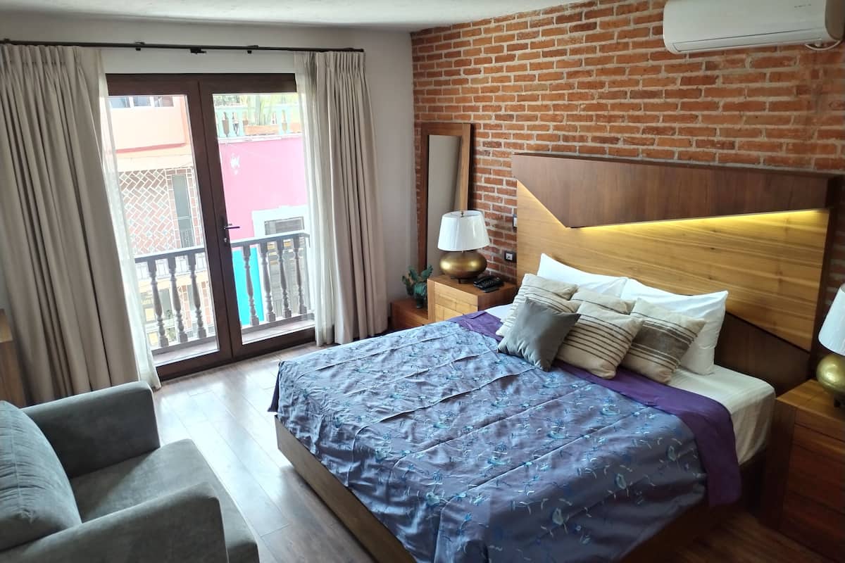 A hotel room with a large bed, a wooden headboard, a nightstand with a lamp, and a view of a red building through a sliding door.