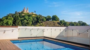 A rectangular swimming pool with a wooden deck surrounding it, set against a backdrop of a clear sky and a hill with a building. - Hotel Casa Chanehque (San Pedro Cholula)