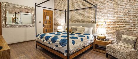 Una cama con dosel con un edredón estampado, una mesita de noche de madera con una lámpara, una silla con un cojín estampado y un espejo con un marco decorado.