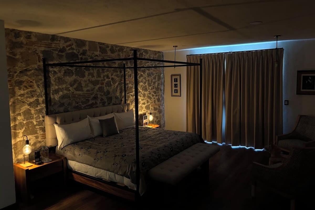 A bedroom with a stone wall, a four-poster bed, a nightstand with a lamp, and a window with curtains.