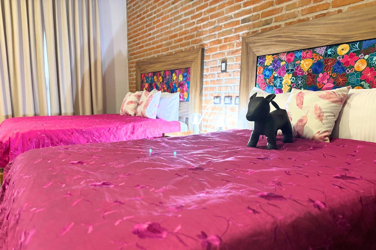 A hotel room with two beds, each covered with a pink bedspread. There is a black dog on the right bed. The room has a brick wall and colorful floral artwork above the beds.
