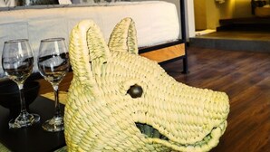 Een kamer met een stenen muur, een bed met een wit hoofdeinde, een houten vloer en een sculptuur van een dierenhoofd, geweven, op een tafel.