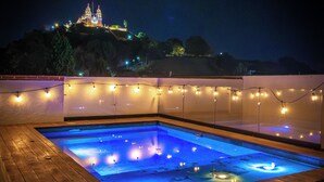 A swimming pool with a wooden deck, surrounded by string lights, and a view of a castle in the background. - Hotel Casa Chanehque (San Pedro Cholula)