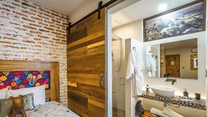 A bedroom with a wooden sliding door, a bed with floral bedding, a colorful abstract painting, and a large mirror reflecting a bathroom with a sink and towel rack. - Hotel Casa Chanehque (San Pedro Cholula)
