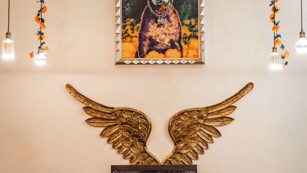 A room with a framed painting of a dog, a bench with a vase of flowers, and a wall-mounted golden wing decoration. - Hotel Casa Chanehque (San Pedro Cholula)
