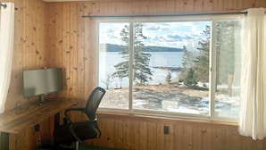 Een kamer met houten wanden, een bureau met een computer en een groot raam met uitzicht op een meer en bomen.