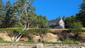 Een huis aan de kust met een steile heuvel, omringd door dichte bomen en een rotsachtig strand.