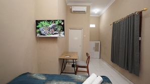 Ein kleiner, kompakter Raum mit einem Bett, einem an der Wand befestigten Fernseher, einem Schreibtisch mit Stuhl und einer Tür zu einem weiteren Raum.
