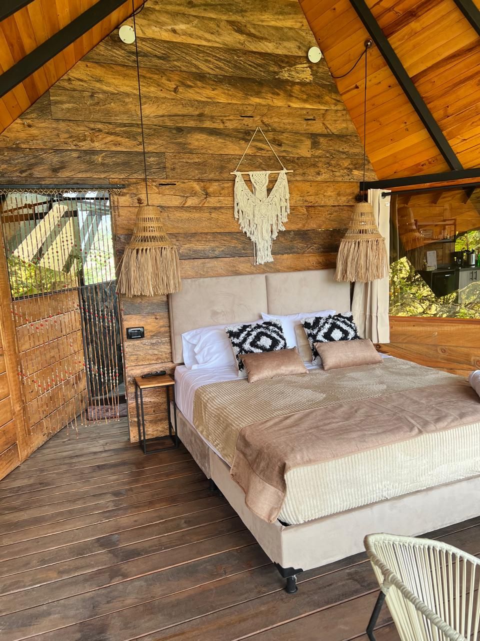 A bedroom with a wooden ceiling, a bed with patterned pillows, a nightstand, and a hanging macrame light fixture.