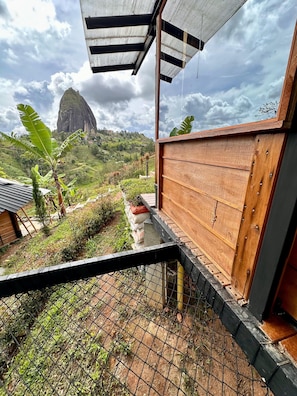 A wooden structure with a metal roof, surrounded by a wire fence, overlooking a lush green landscape with a prominent mountain in the distance. - Ecoedén Glamping (Guatapé)