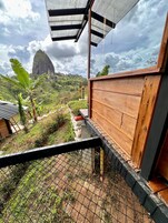 Una estructura de madera con techo metálico, rodeada por una cerca de alambre, con vistas a un paisaje verde exuberante y una montaña destacada en la distancia.