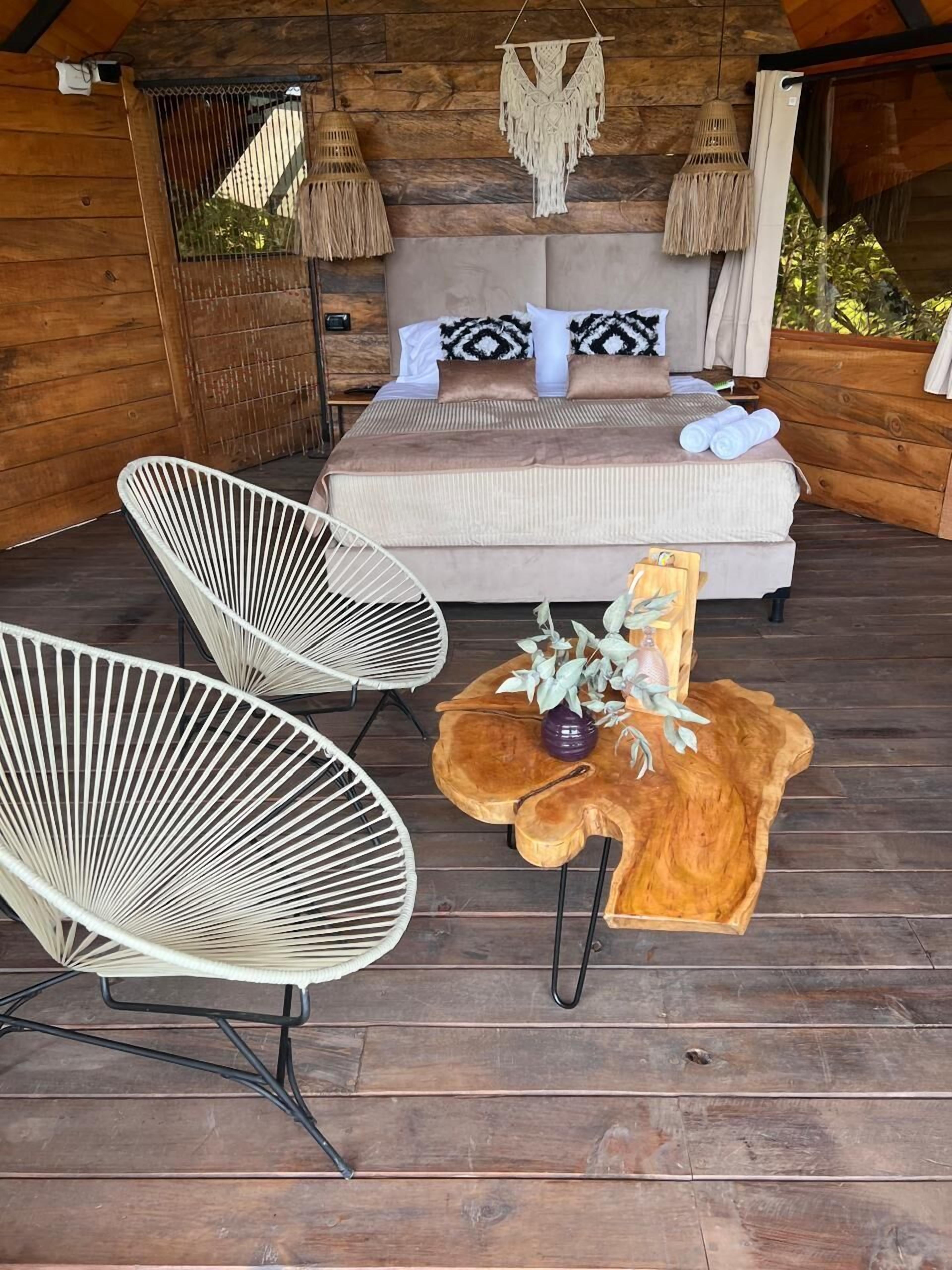 A wooden deck with a bed, two white chairs, and a wooden table with a vase and plants.