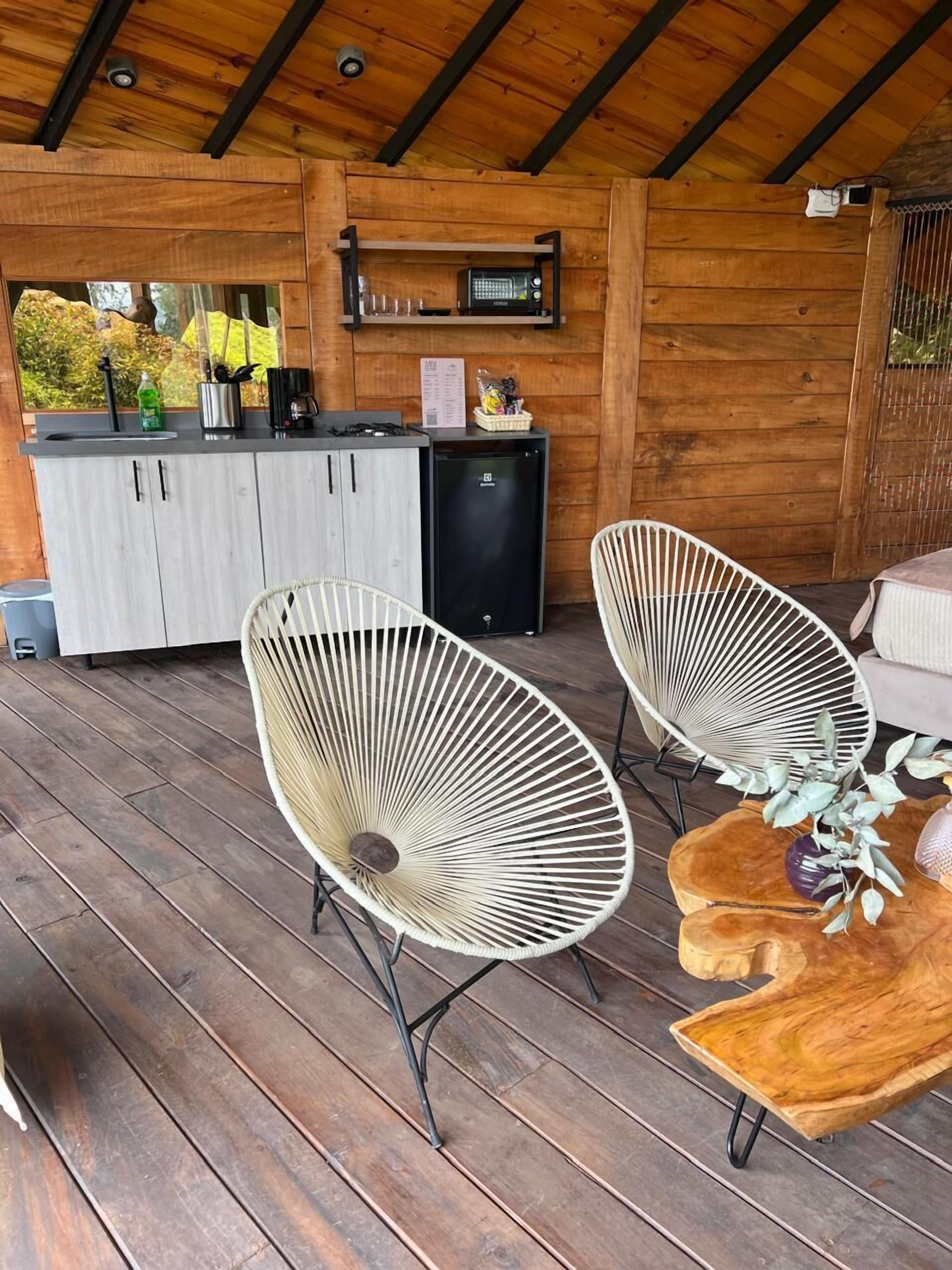 A wooden deck with two white woven chairs, a small table with decorative items, and a kitchenette area with cabinets and a refrigerator.