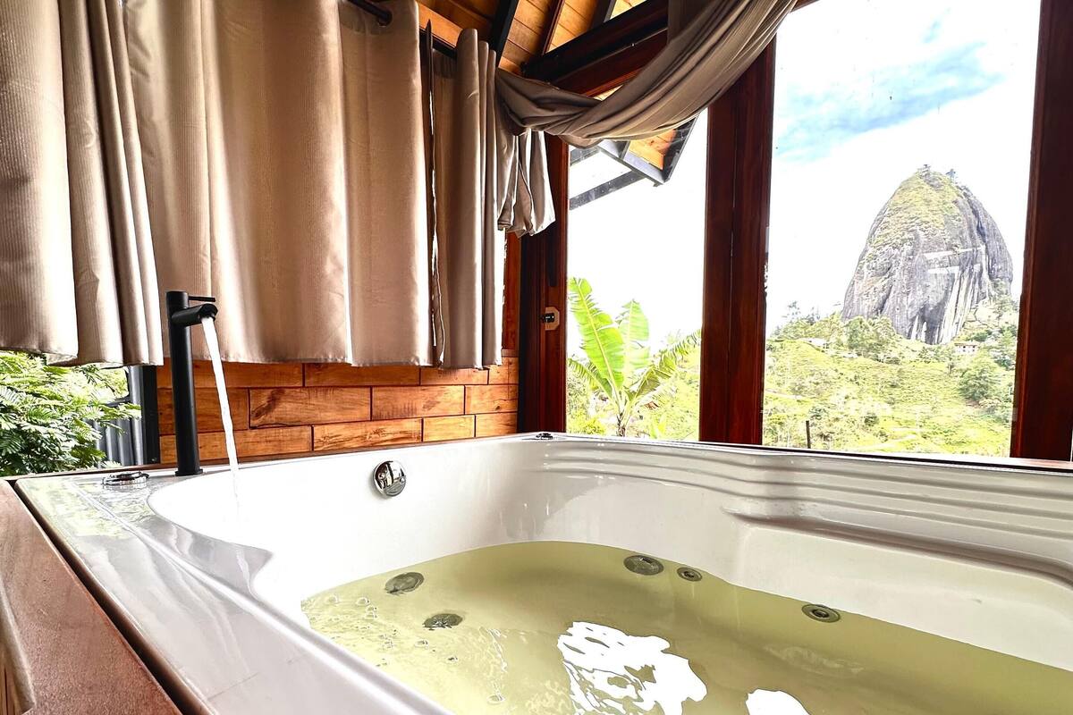 A hot tub in a room with wooden beams and large windows overlooking a landscape with a prominent rock formation.