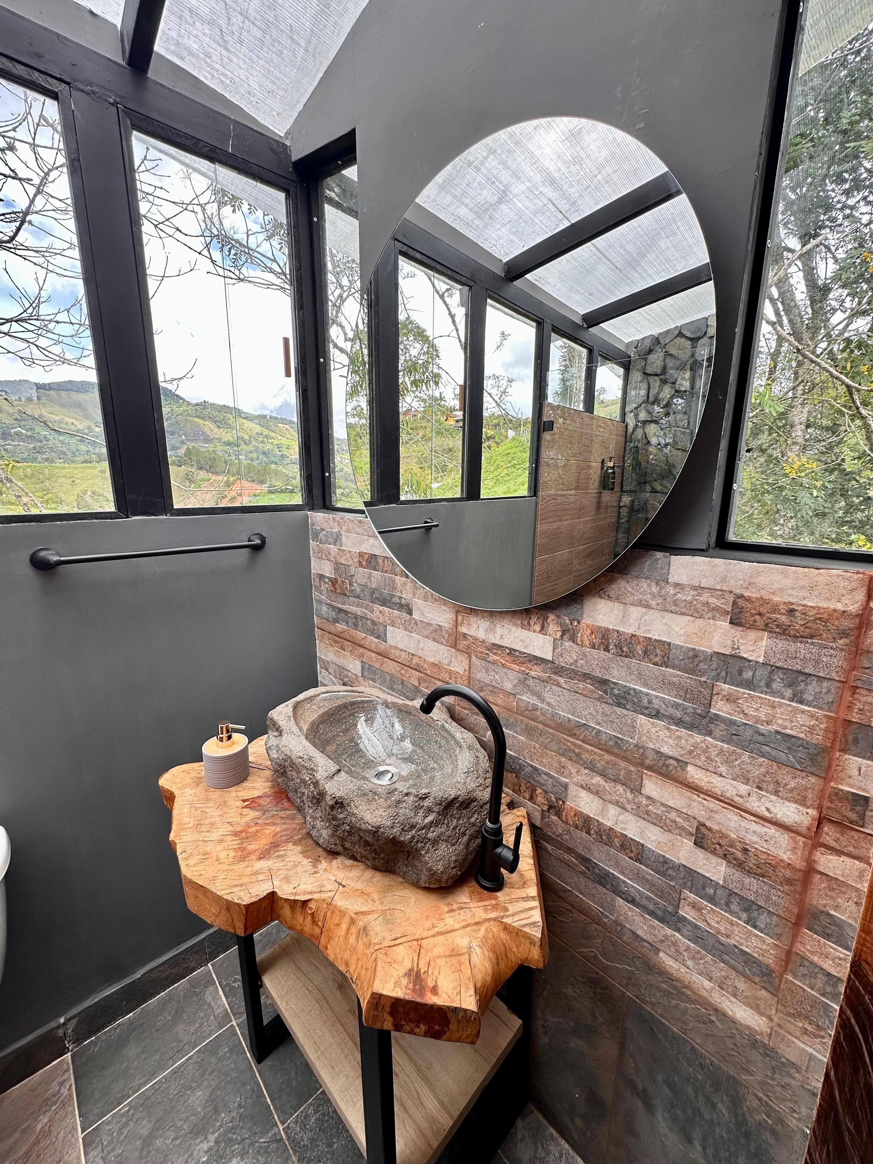 Ett badrum med ett handfat i sten, ett träbord och en stor spegel som reflekterar omgivningen utomhus.