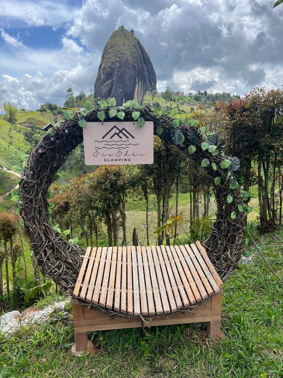 A wooden bench with a sign above it, surrounded by greenery and a mountainous backdrop.