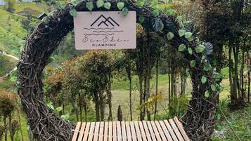 A wooden bench with a sign above it, surrounded by greenery and a mountainous backdrop.