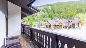 Un terrazzo in legno con mobili in vimini, affacciato su un lodge dalla facciata in pietra e vista su un pendio boscoso.