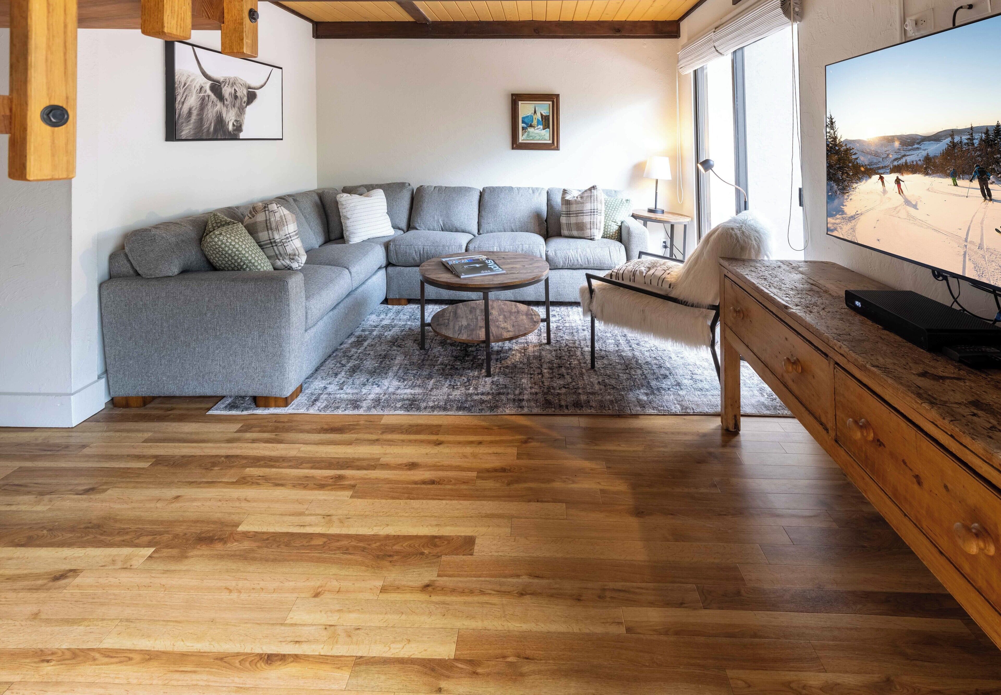A living room with a grey sectional sofa, a round coffee table, a wooden cabinet, and a television displaying a snowy landscape.