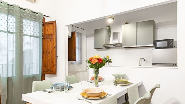 Un coin repas avec une table dressée pour quatre personnes, un vase rempli de fleurs et une cuisine équipée d’appareils modernes.