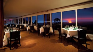 Un restaurant avec des tables dressées pour le dîner, de grandes fenêtres et une vue sur le coucher de soleil.