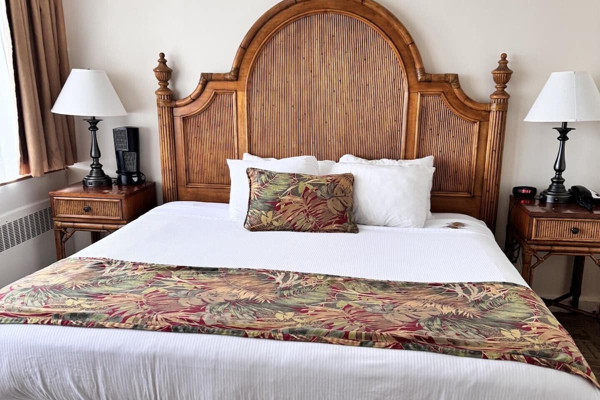 A hotel room with a large bed, wooden headboard, two bedside tables with lamps, and a window with curtains.