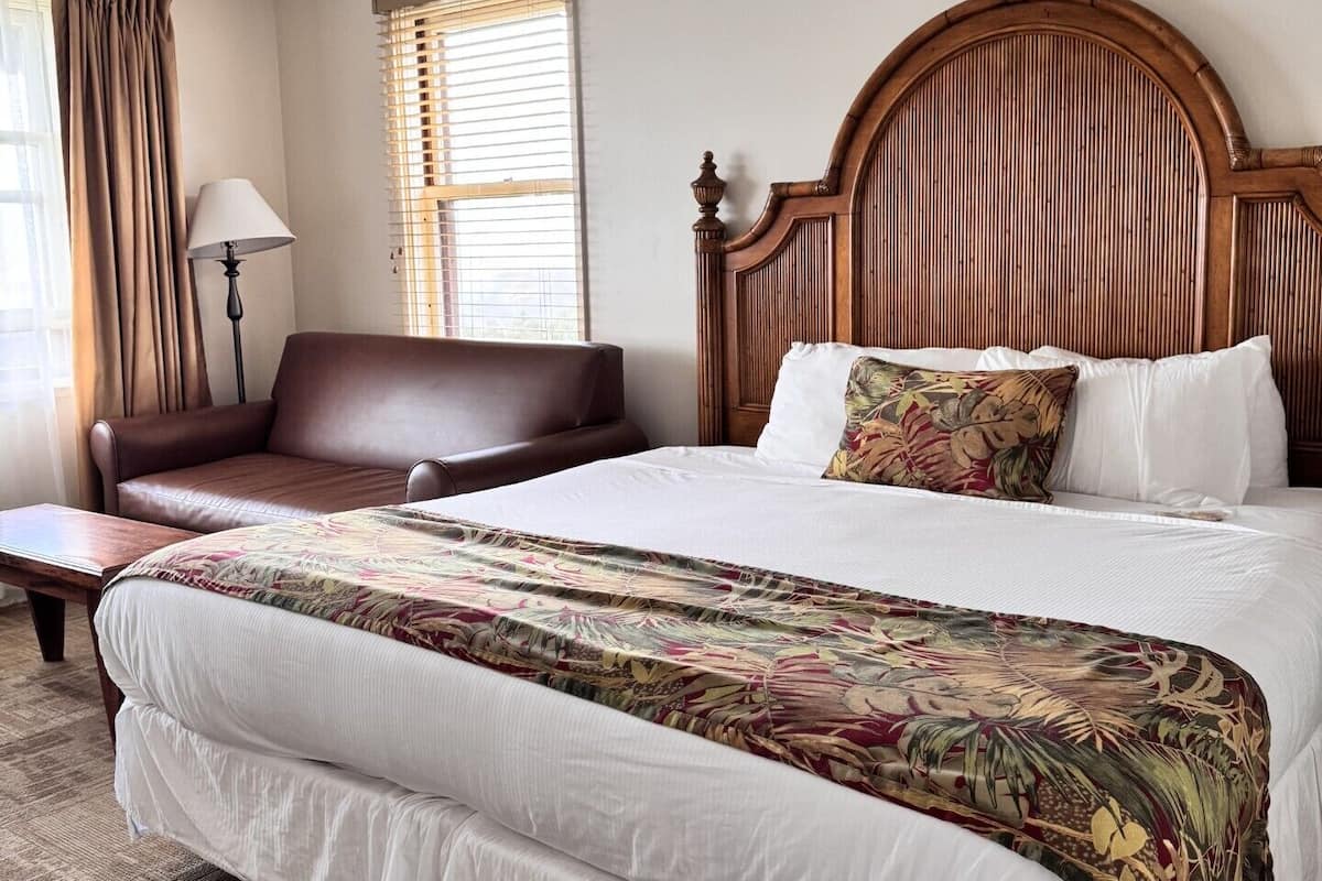 A hotel room with a large bed, a wooden headboard, a brown leather sofa, a bedside table, and a lamp.