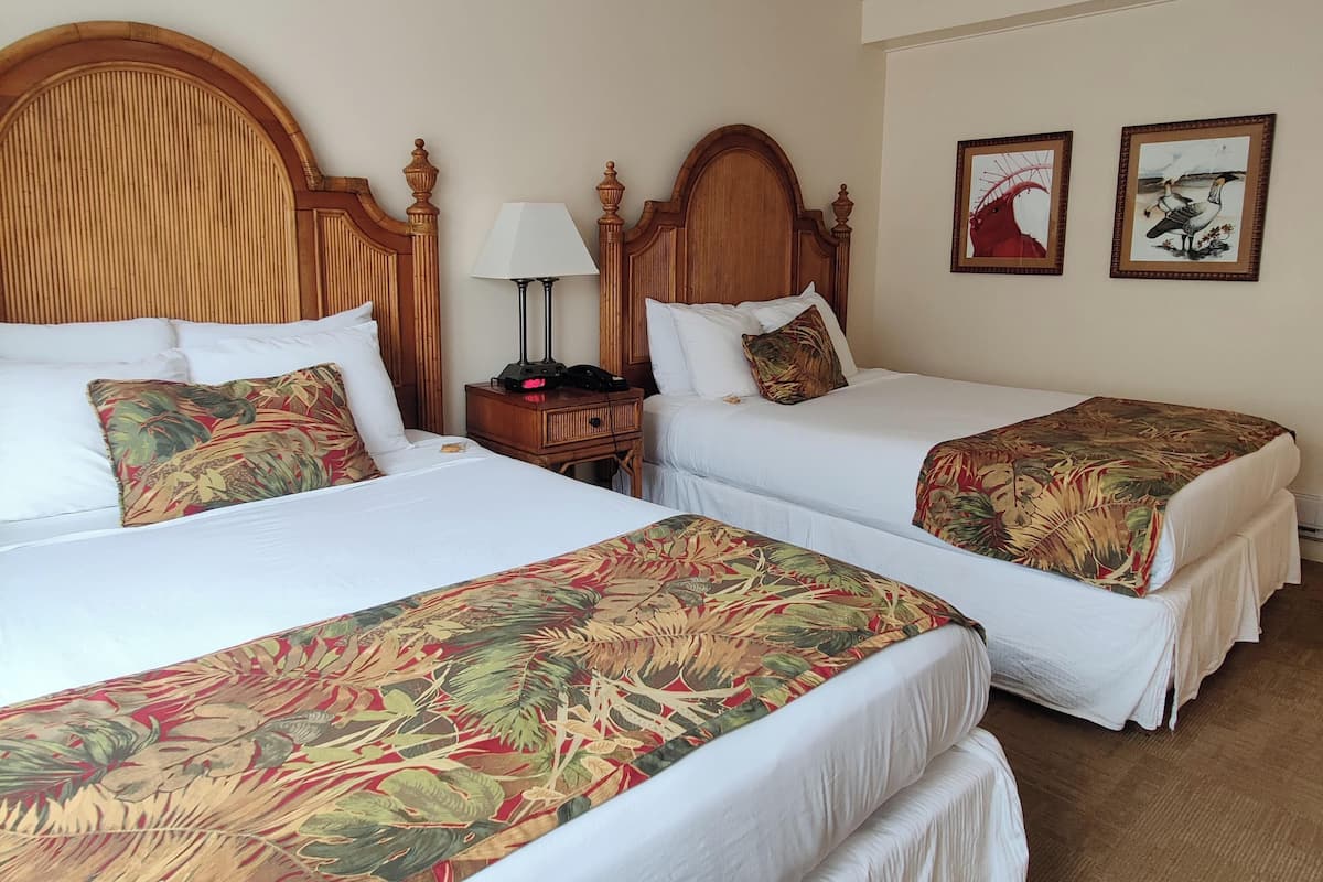 A hotel room with two beds, each with a wooden headboard and white bedding. There is a nightstand with a lamp and a small table between the beds. The room has a simple, clean design with a wooden floor.