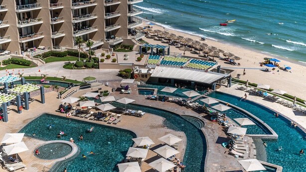 Un complexe hôtelier doté de plusieurs piscines, d’une plage et d’un immeuble de grande hauteur avec des balcons.