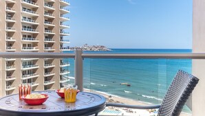 Un balcon avec une table dressée pour deux personnes, offrant une vue sur une plage et sur les bâtiments d’une ville lointaine.