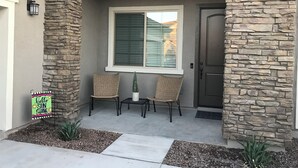 A house entrance with stone walls, a window with blinds, two wicker chairs, a small potted plant, and a 'hello sun shine' sign. - Cozy Casa Bliss: 3 bedroom Desert Home (Casa Grande)