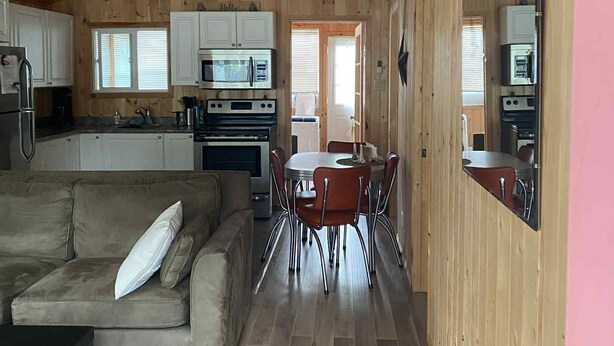 A spacious living area with a wooden ceiling, a ceiling fan, a sofa, a dining table with chairs, and a kitchen area with cabinets and appliances. - Cozy Cabin in Grand Beach.  (Grand Marais)
