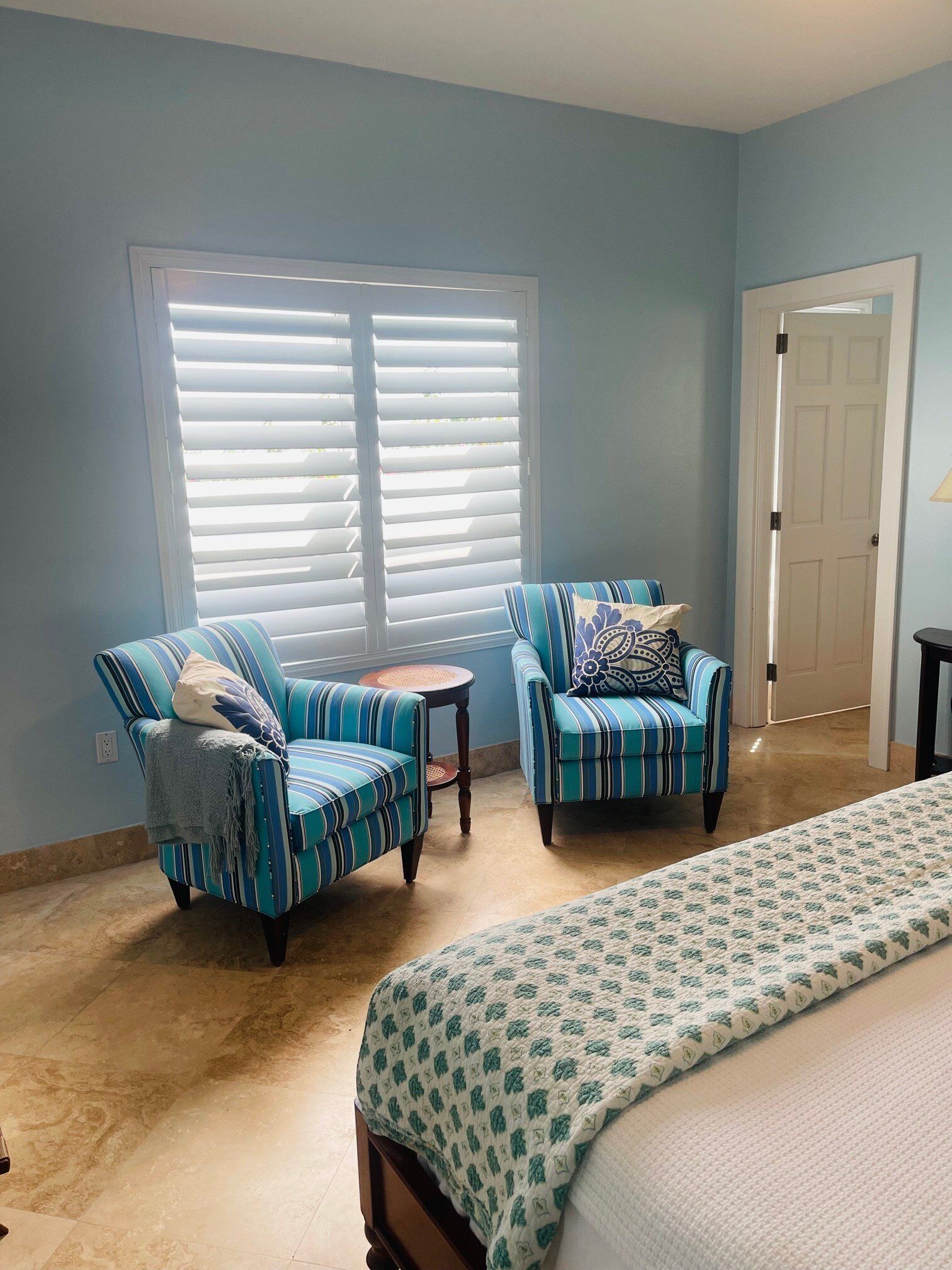 A room with two striped armchairs, a small round table, a bed with a patterned bedspread, and a window with blinds.