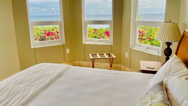 Un dormitorio con cama, mesita de noche, lámpara y tres ventanas con vistas a flores y al mar.