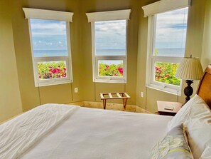 Ein Schlafzimmer mit einem Bett, einem Nachttisch, einer Lampe und drei Fenstern mit Blick auf Blumen und das Meer.