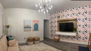 A living room with a beige sofa, a patterned rug, a TV on a wooden shelf, and a wall with a leaf-patterned wallpaper. - Chic 1-bedroom 1-bath house with AC and WiFi in the heart of Atlanta (Atlanta)