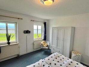 Une chambre avec un lit, une armoire, une chaise, un radiateur, une fenêtre donnant sur la mer et une plante en pot.
