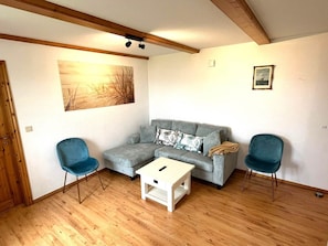 Un salon chaleureux avec un canapé gris, deux fauteuils bleus, une table basse blanche, un plancher en bois, un tableau au mur et une photographie encadrée.