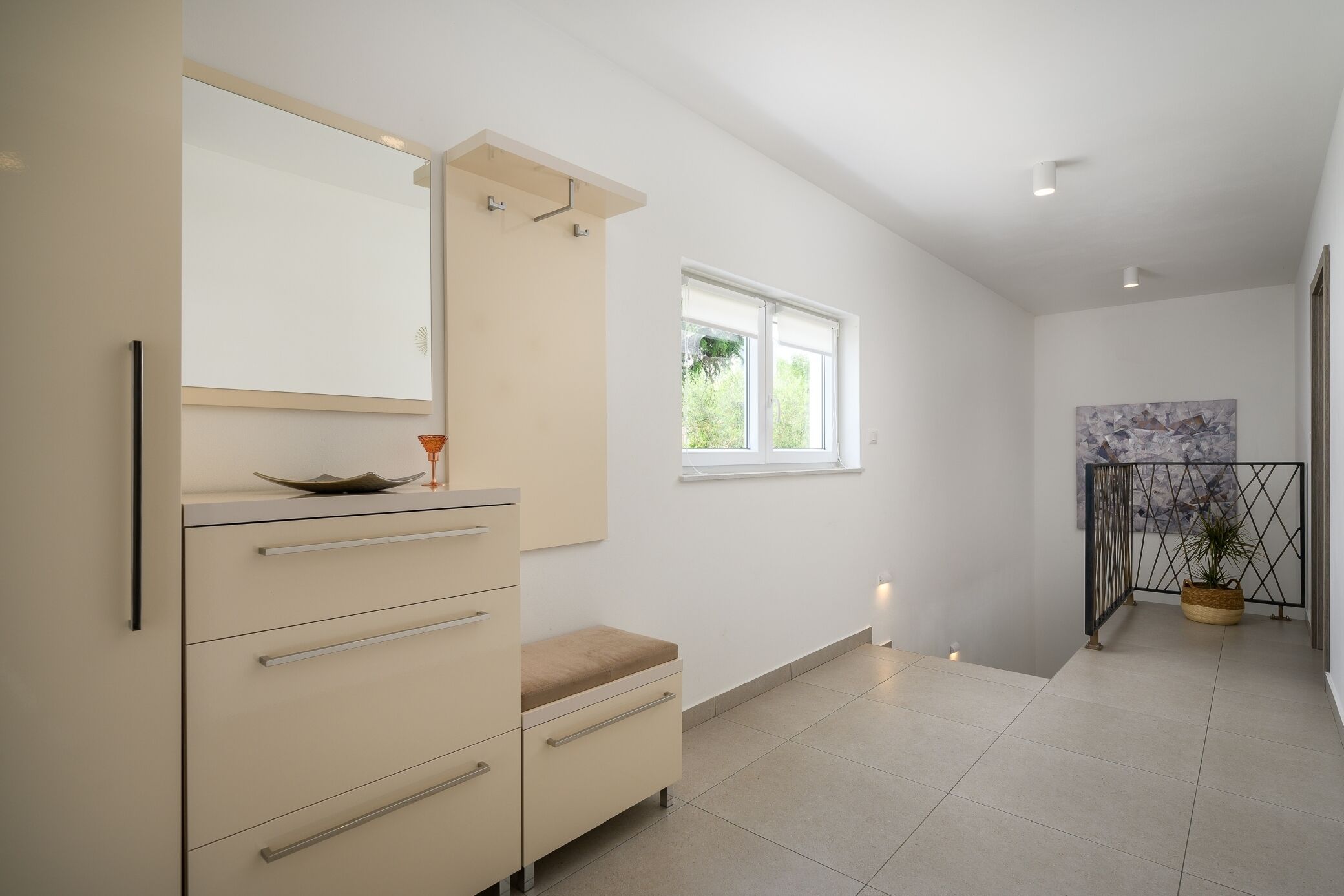 A modern bathroom with a large mirror, a vanity with drawers, a bench, and a window providing natural light.