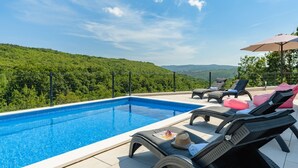 Une piscine entourée d’une clôture et de verdure, avec des chaises longues et un parasol.