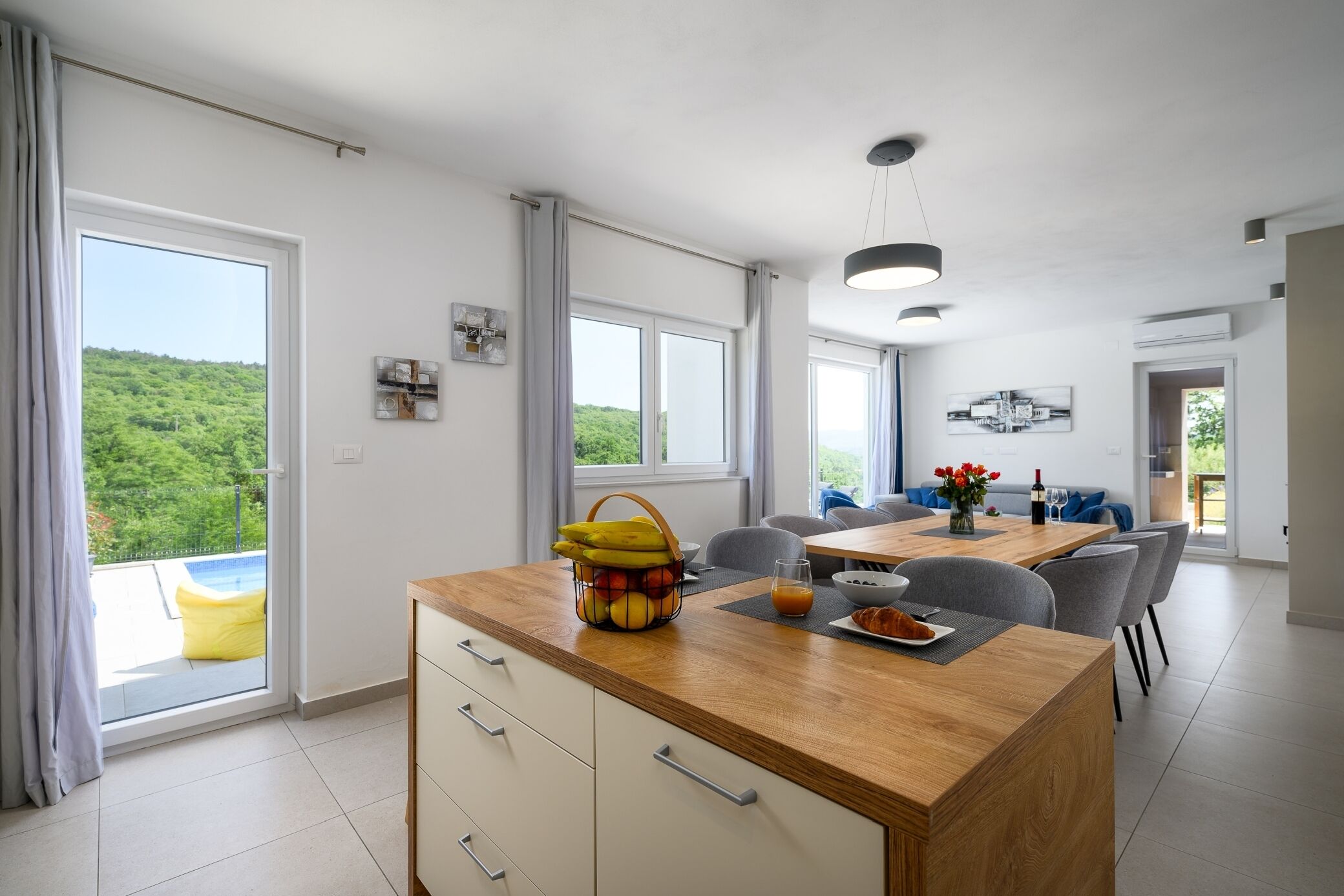 A modern kitchen with a wooden countertop, a dining area with a table and chairs, and large windows offering a view of greenery.