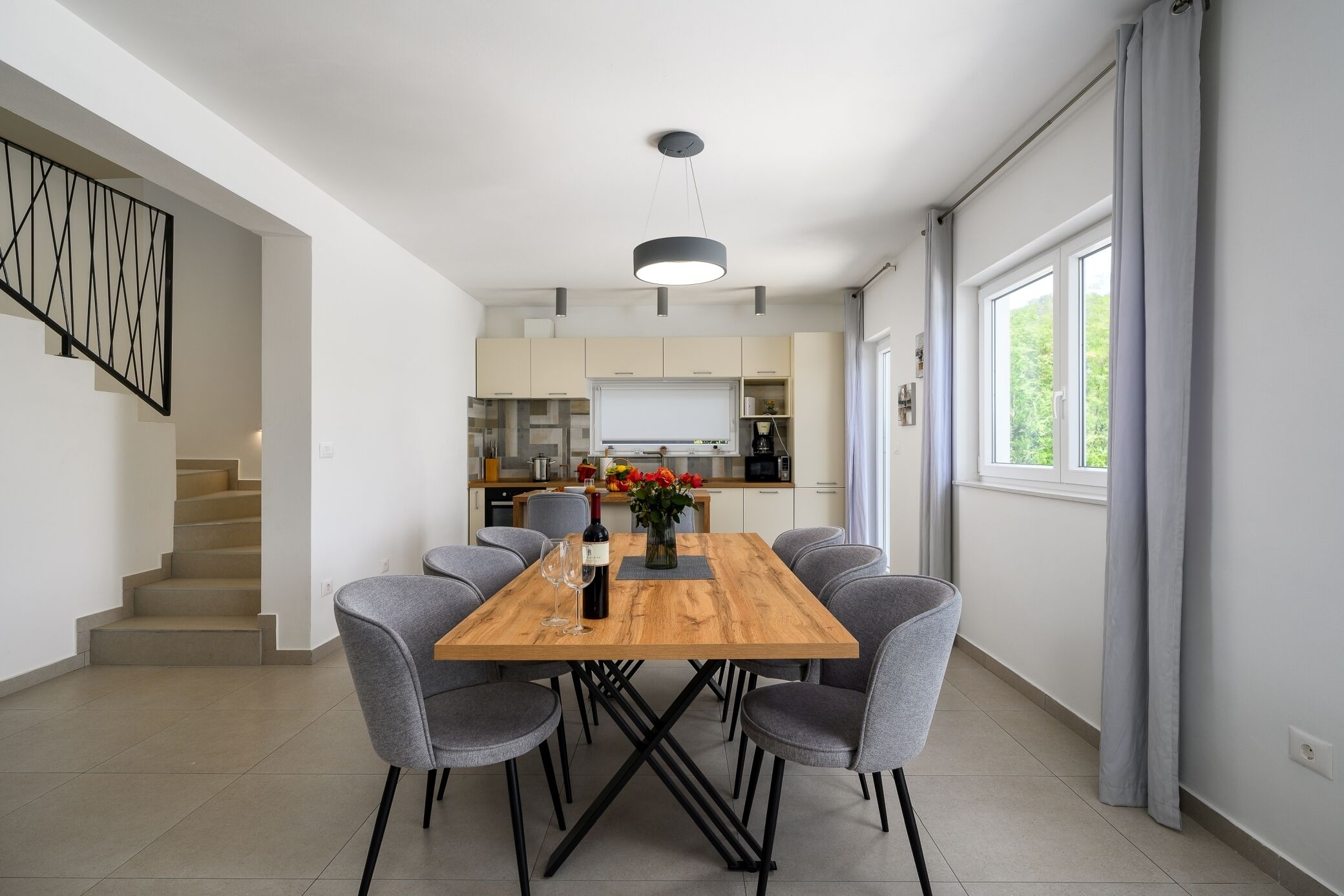 Een moderne eetruimte met een houten tafel en grijze stoelen, een trap aan de linkerkant en een keuken op de achtergrond.