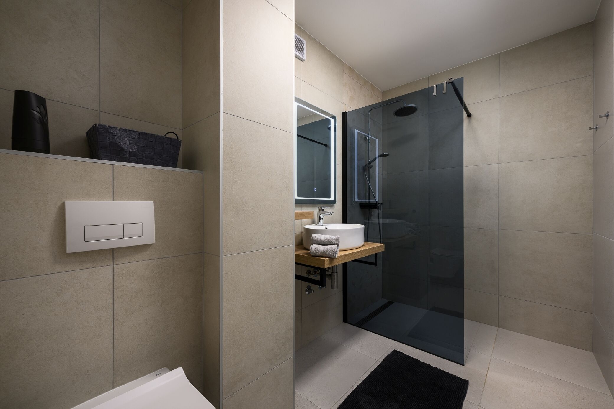 A modern bathroom with a glass-enclosed shower, a white sink with a wooden countertop, and a black rug on the floor.