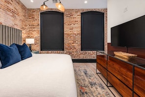 A bedroom with a brick wall, a bed with blue pillows, a wooden dresser, a flat-screen TV, and two windows with black shutters.
