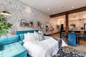 Un dormitorio moderno con un sofá azul verdoso, una cama grande y una cocina al fondo.