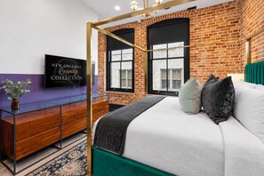 A bedroom with a brick wall, a large bed with pillows, a TV on the wall, and a wooden dresser.