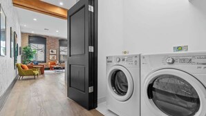 A laundry room with two front-loading washing machines, a wooden floor, and a door leading to a living area with a sofa and chairs. - Modern Comfort Suite Pool Gym Luggage Lockers Hotel (New Orleans)