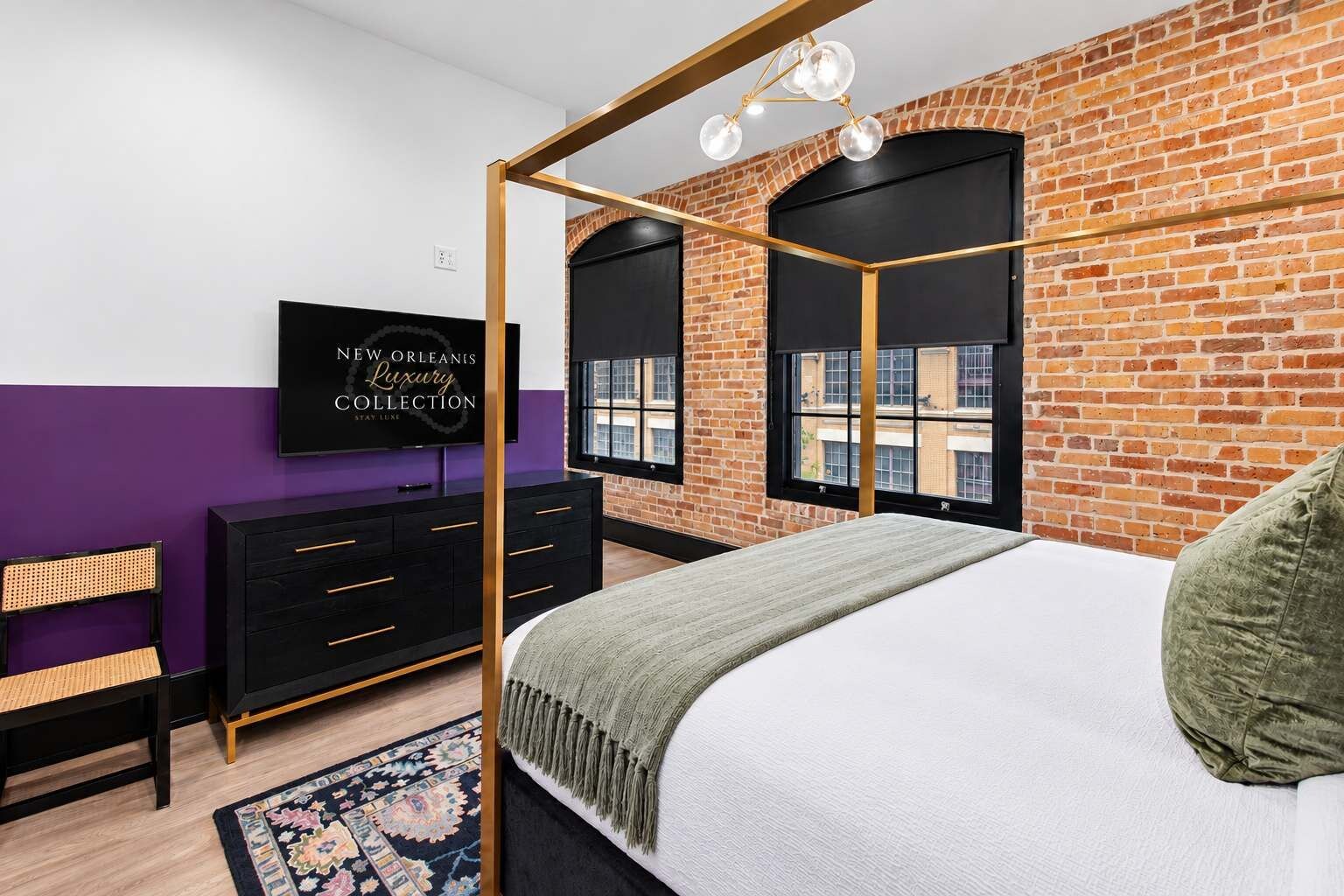 Un dormitorio moderno con pared de ladrillo, un armario negro, una cama con almohada verde y un televisor mostrando 'NEW ORLEANS' LUXURY COLLECTION.